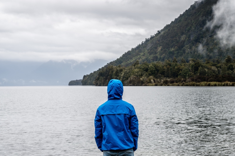 A man wearing a blue rain jacket faces a rainy ocean landscape
